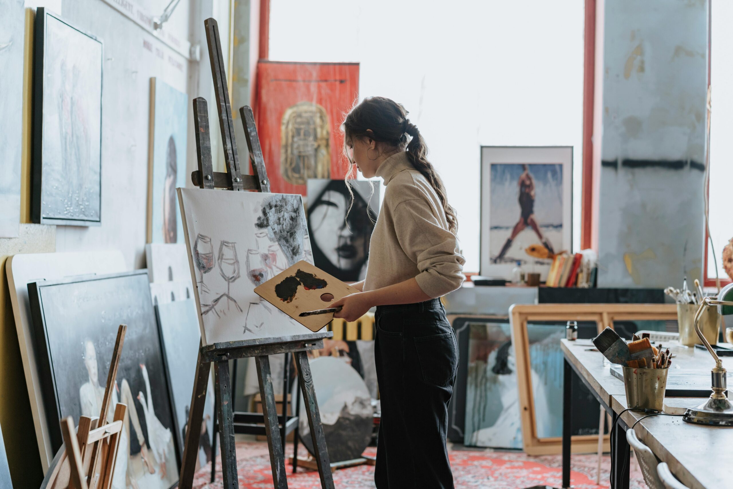 A woman artist works on a painting in a creative studio space filled with art and light.